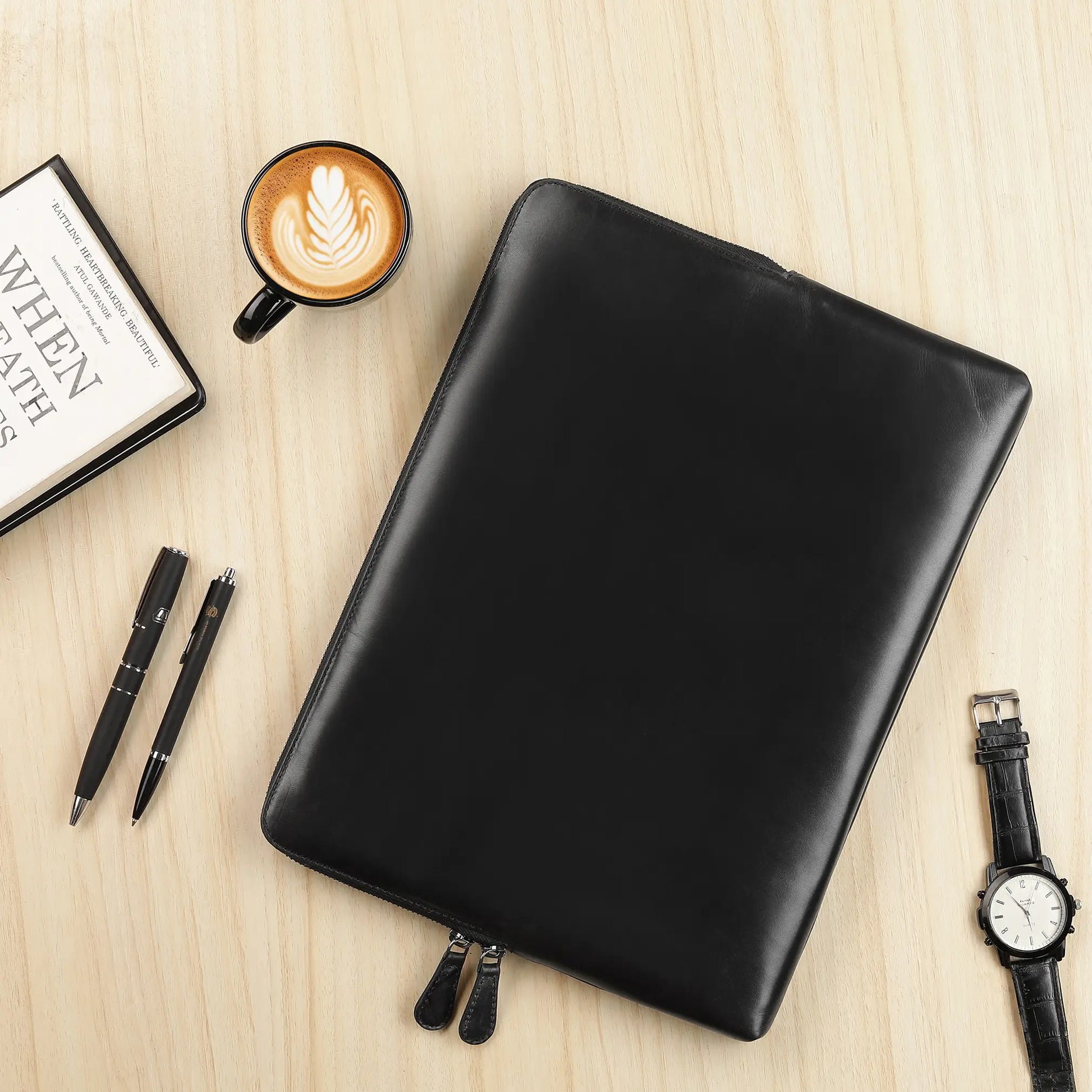 Black leather laptop sleeve on a wooden surface with a coffee cup, book, pens, and watch.