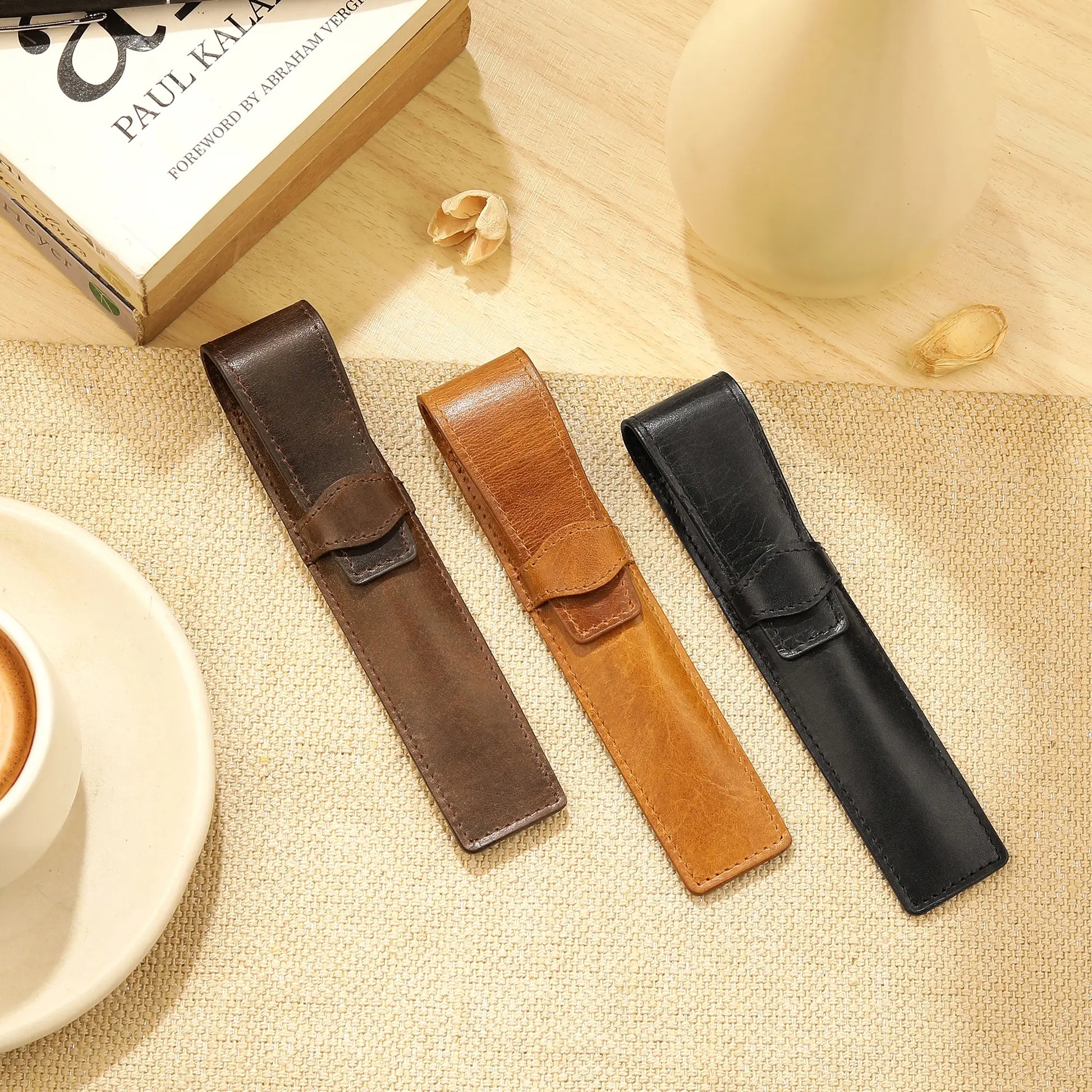Three leather pen holders in brown, black, and tan on a textured surface with a book and cup in the background.