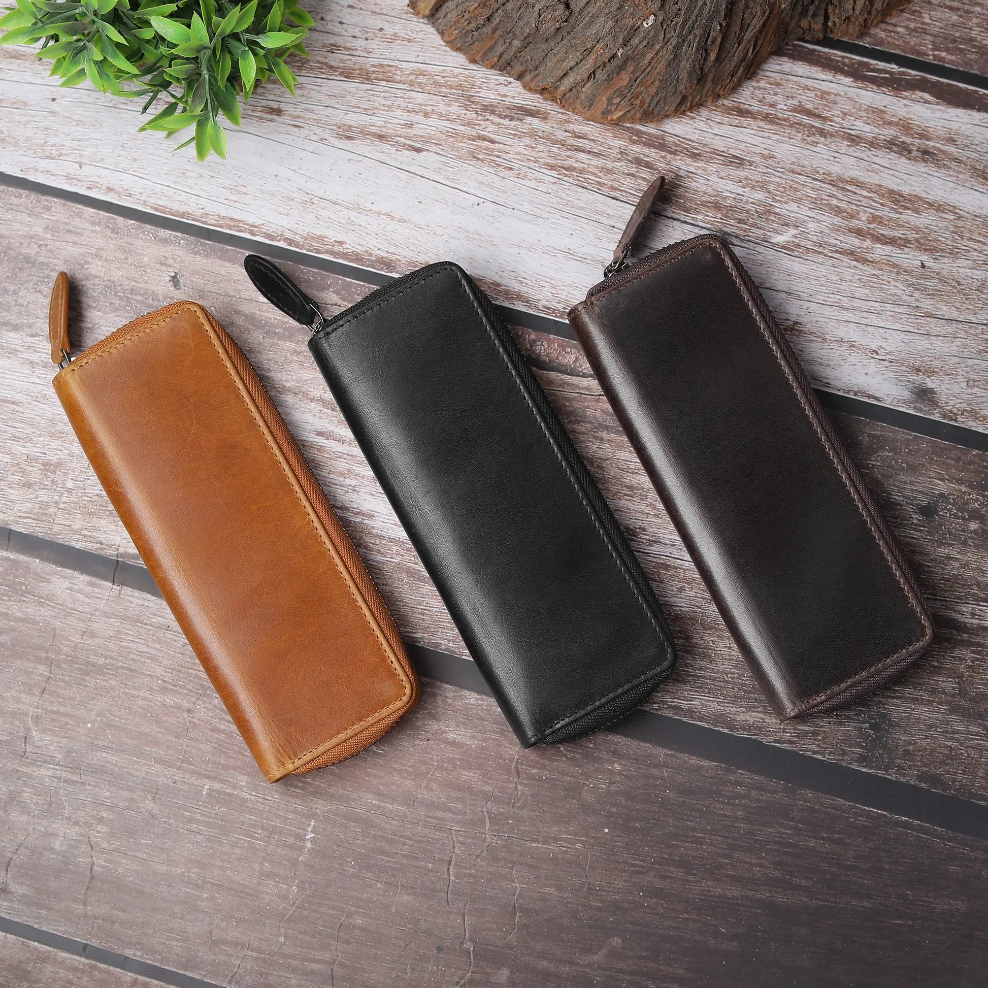 Three leather pen case in brown, black, and dark brown on a wooden surface.