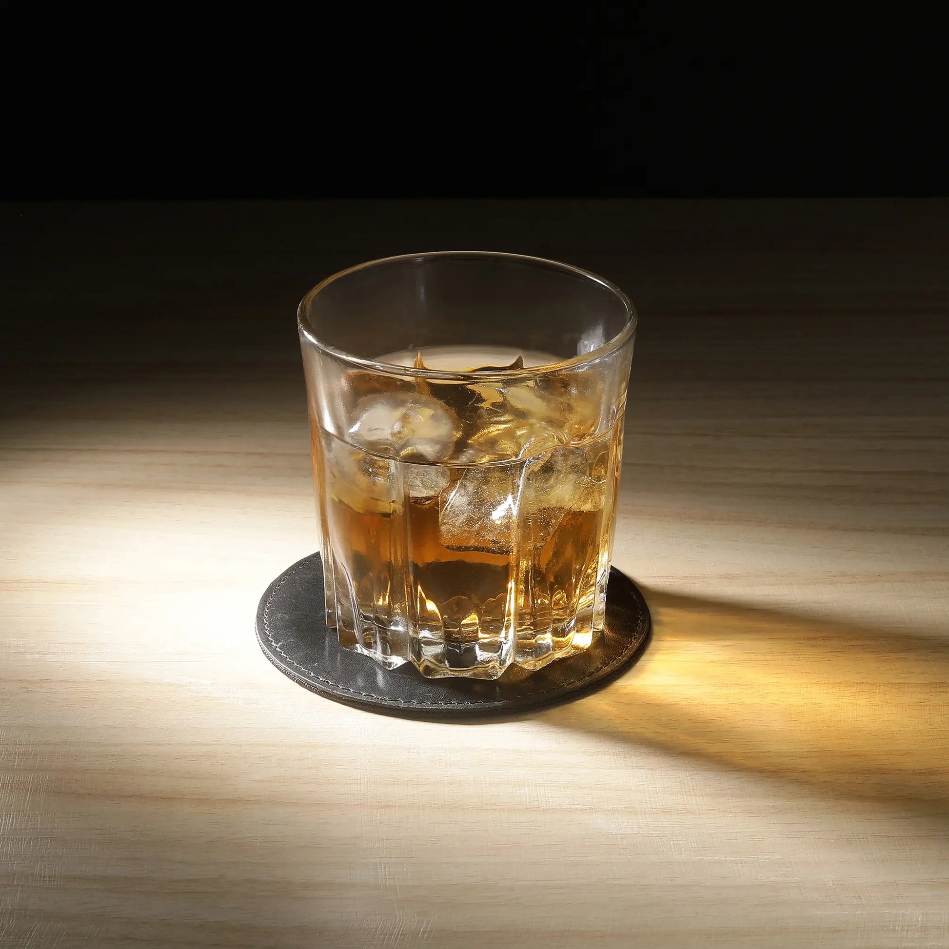 Glass of whiskey with ice on a black leather coaster on a wooden table