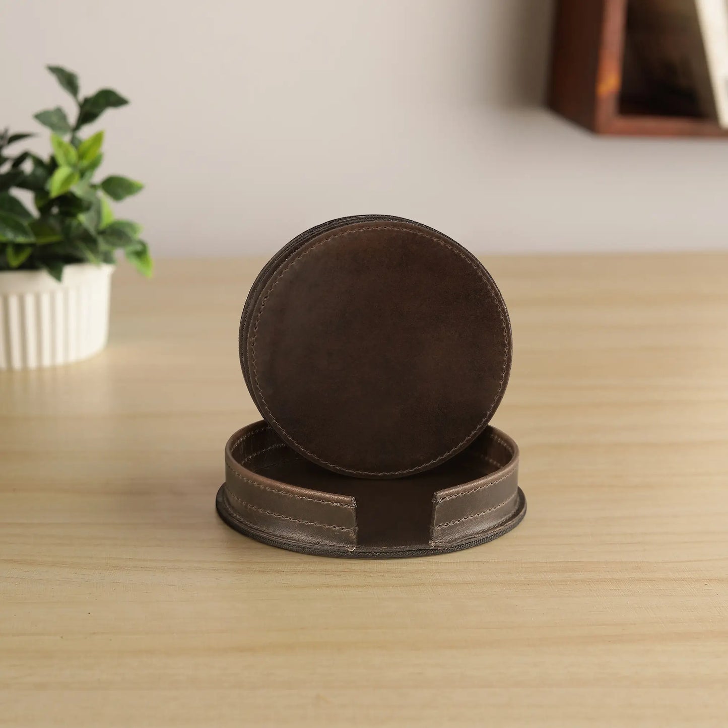 Two brown leather coasters on a wooden surface with a plant in the background.