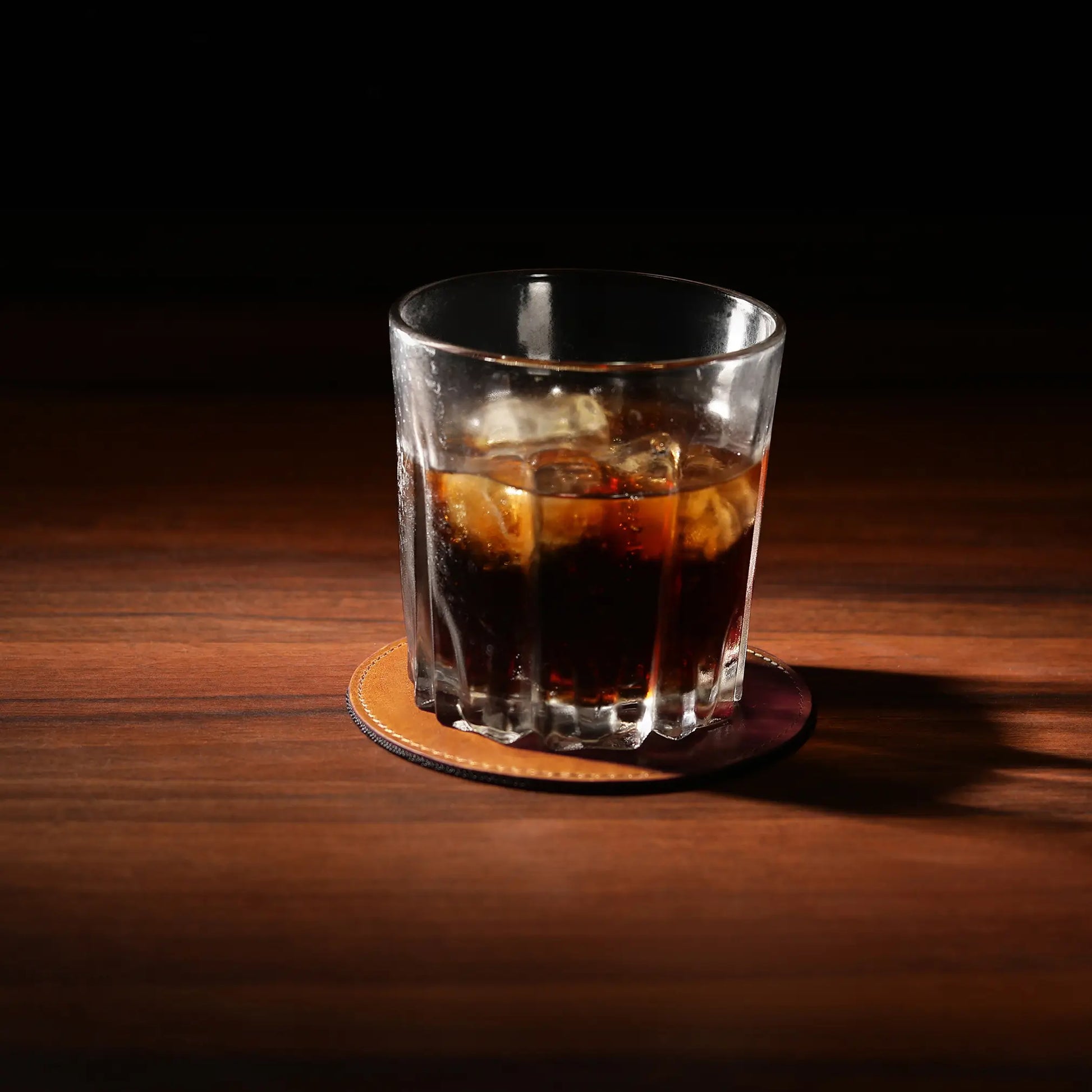 Glass of dark liquid with ice on a tan leather  coaster against a dark background