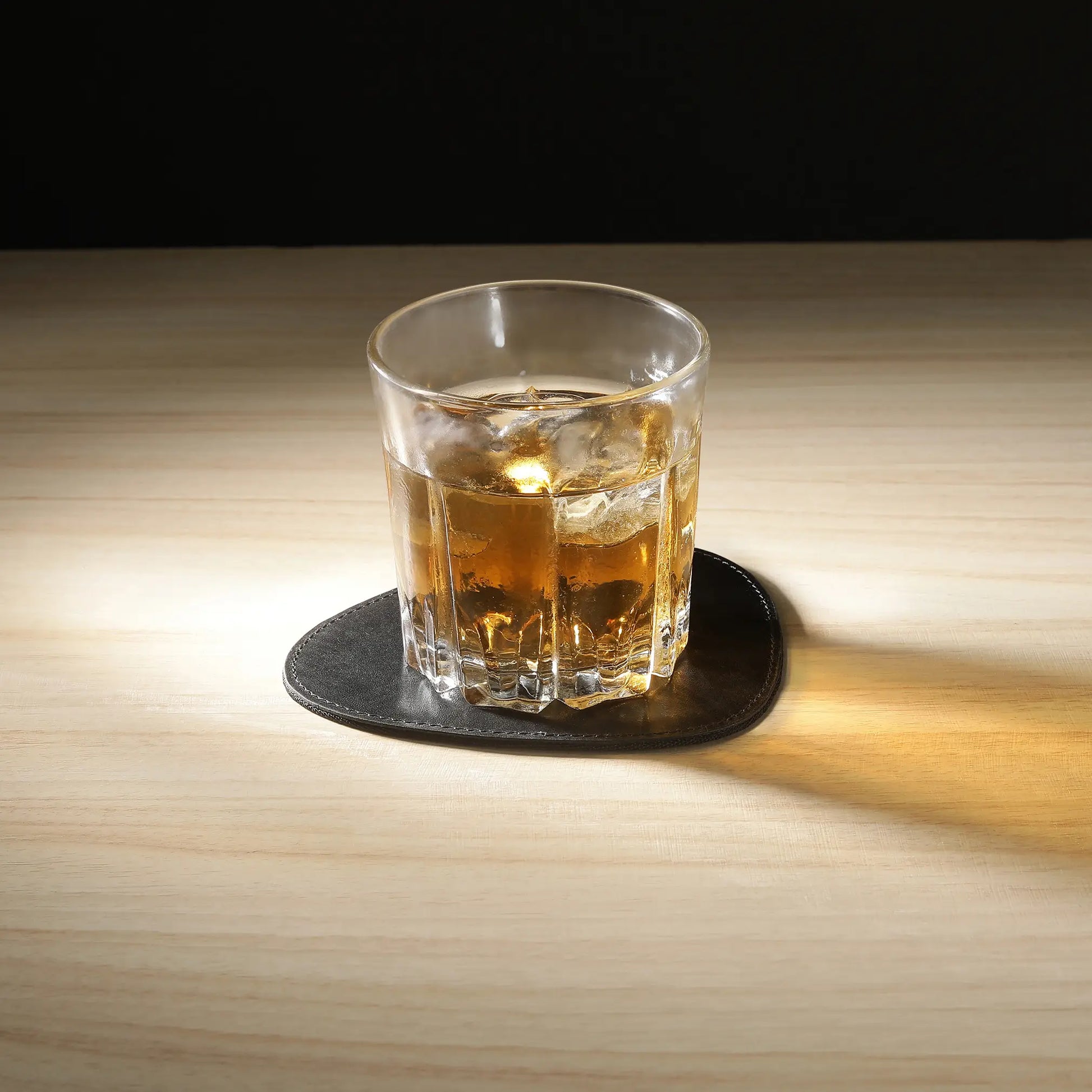 Glass of whiskey with ice on a black coaster on a wooden table