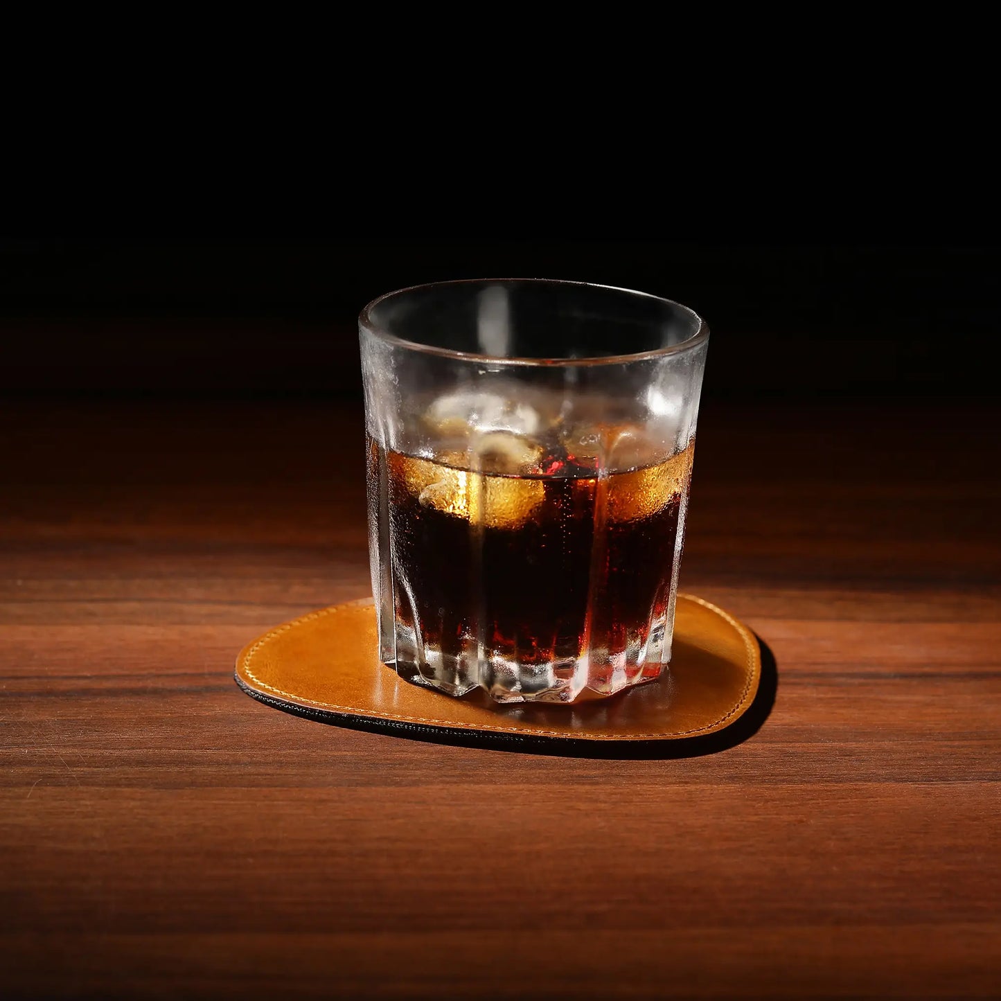 Glass of dark liquid with ice on a wooden leather tan coaster against a dark background