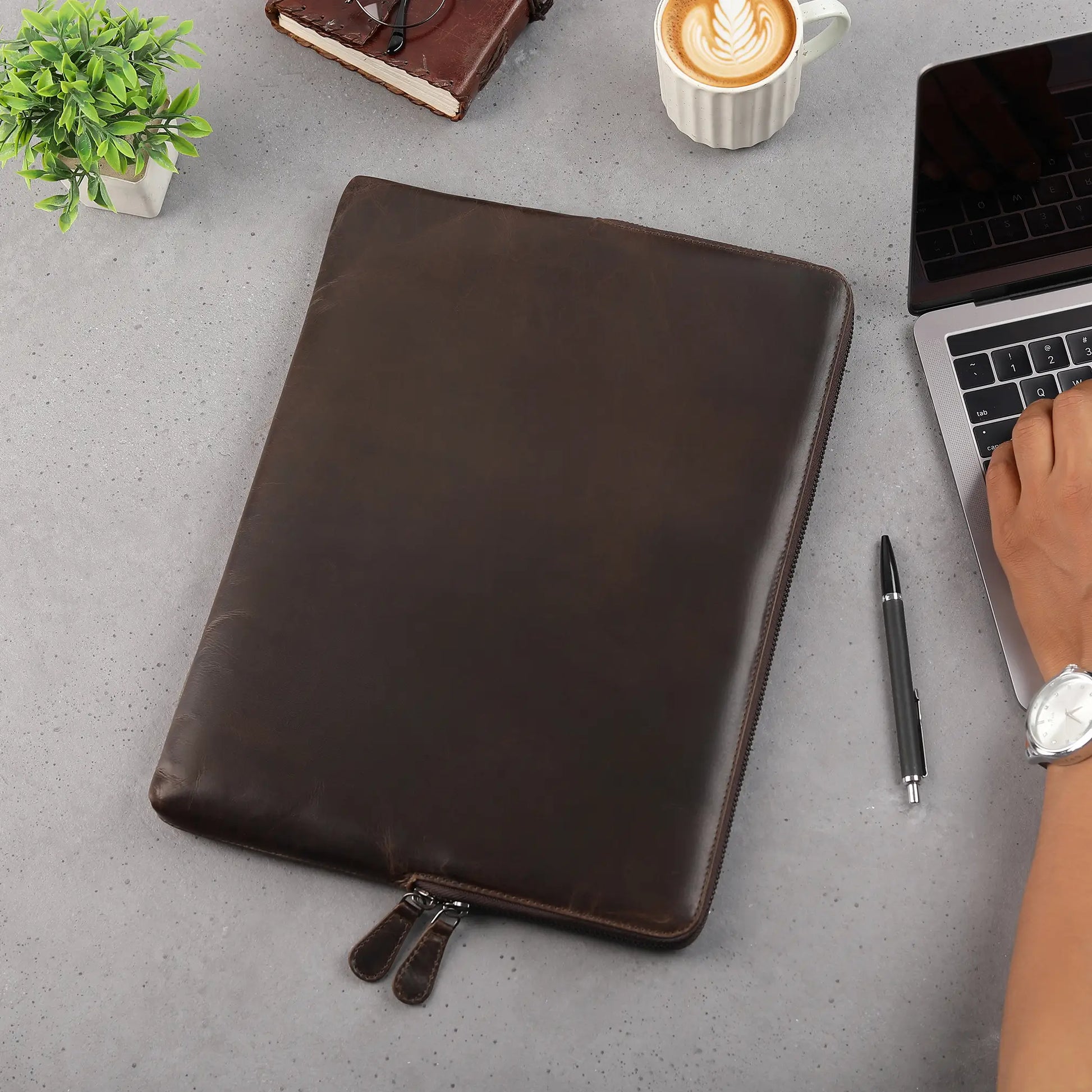 Brown leather laptop sleeve on a desk with a laptop, pen, and coffee cup.