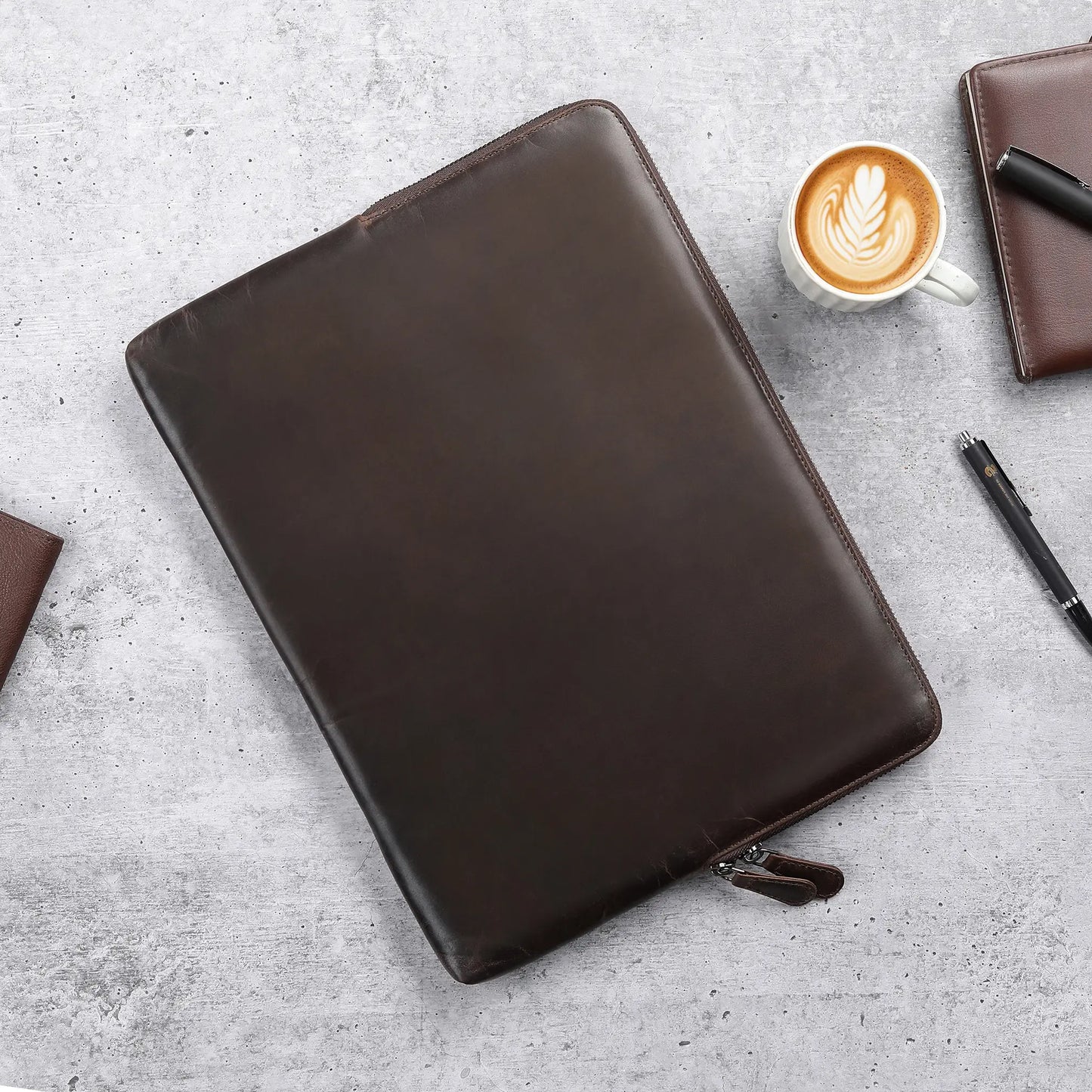 Brown leather zippered laptop sleeve  on a gray surface with a cup of coffee and pen.