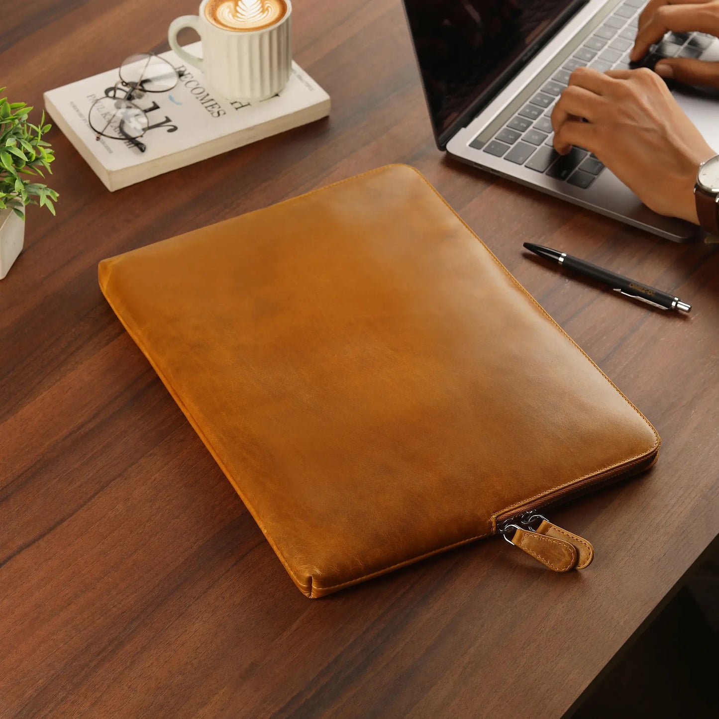 Tan leather laptop sleeve on a wooden desk with a person using a laptop.