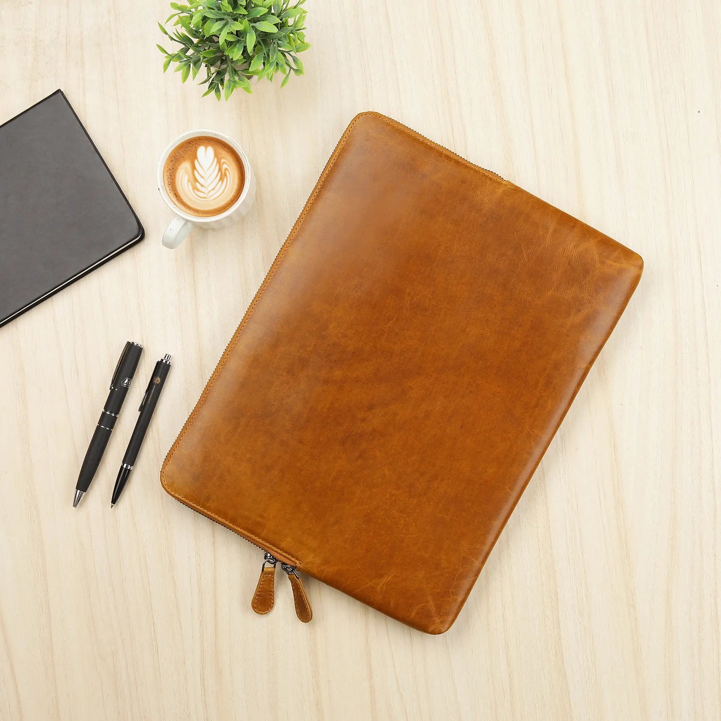 Tan leather laptop sleeve on a wooden surface with a cup of coffee, pen, and plant.