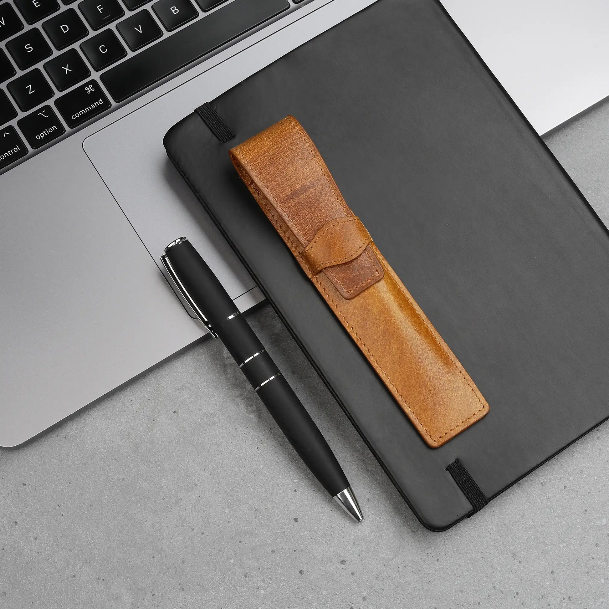 Black pen with tan leather pen holder on a gray surface next to a laptop.