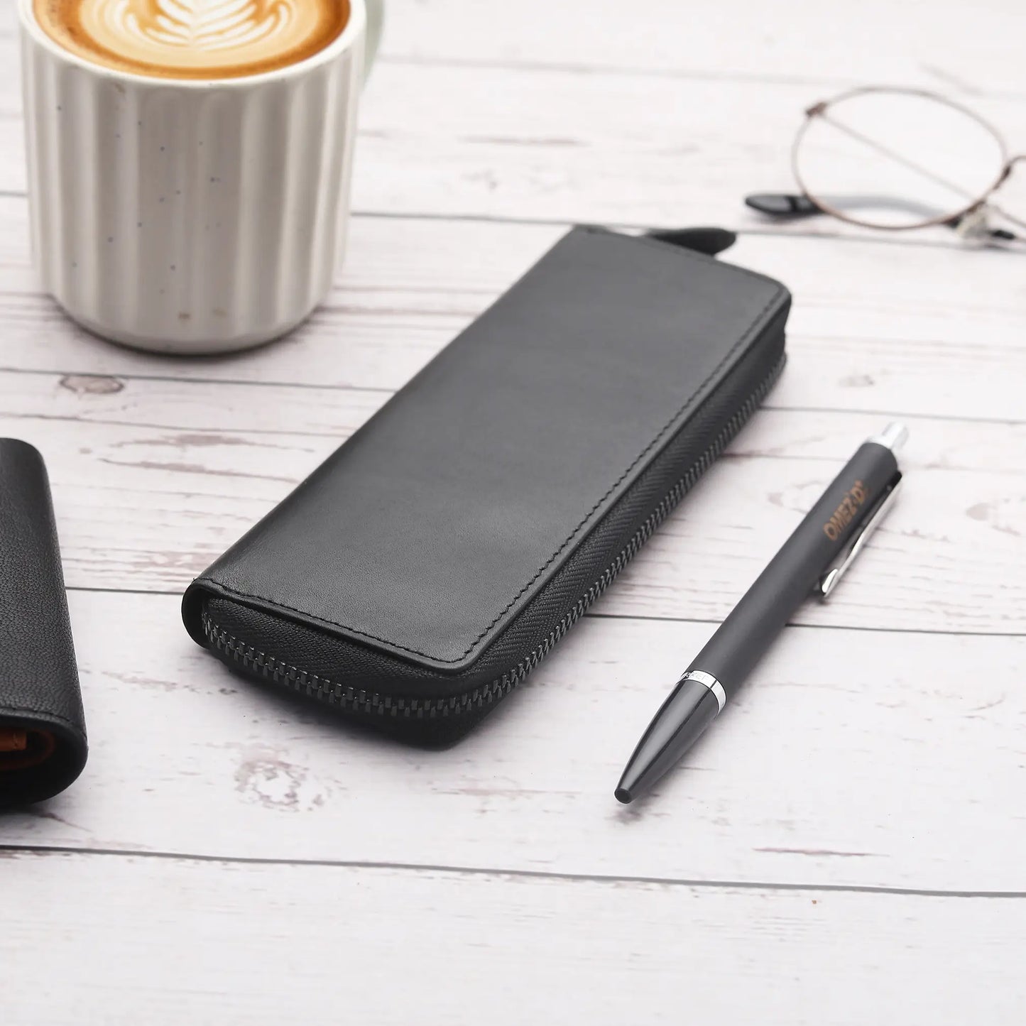 Black leather pen case, pen, and glasses on a wooden surface with a cup of coffee.
