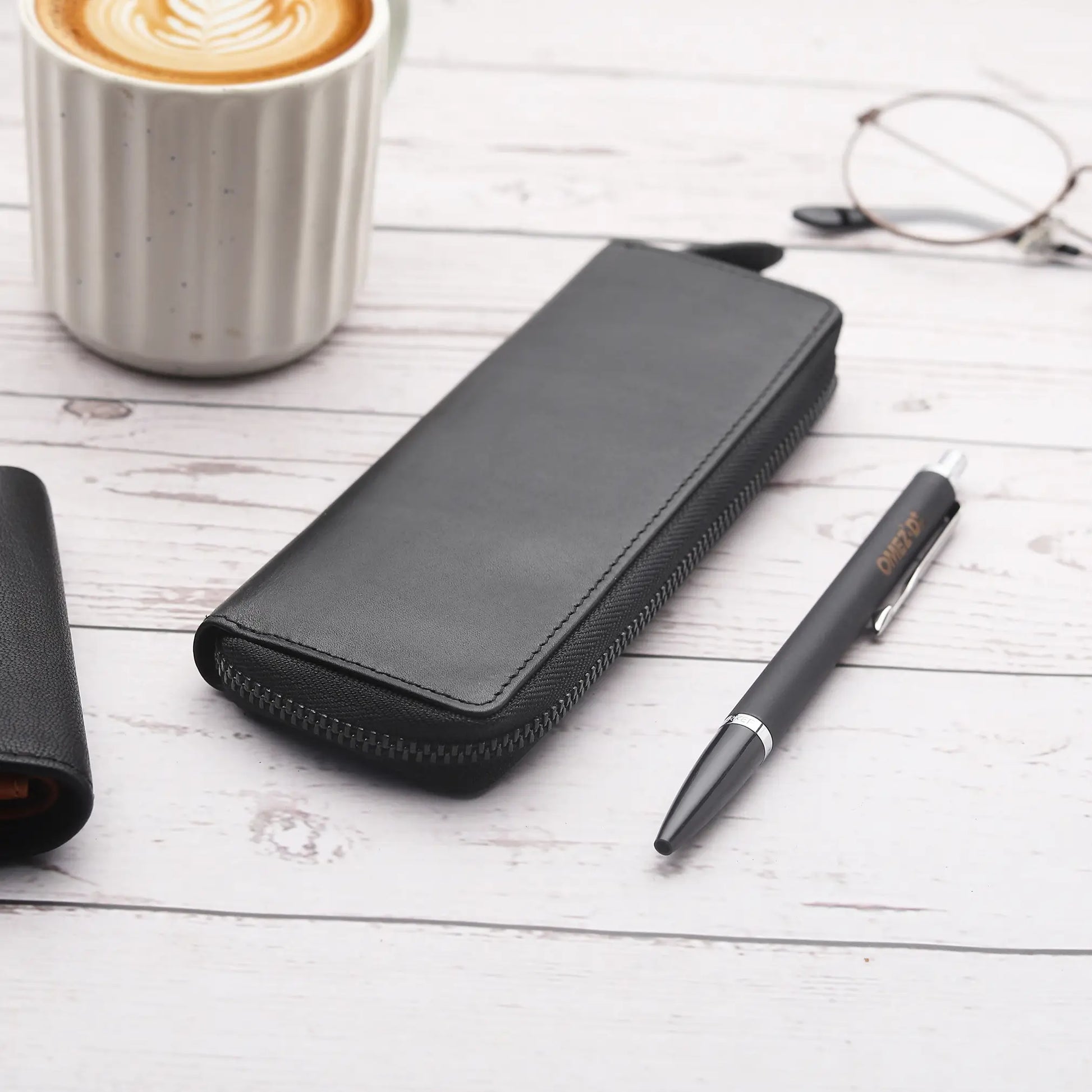 Black leather pen case, pen, and glasses on a wooden surface with a cup of coffee.