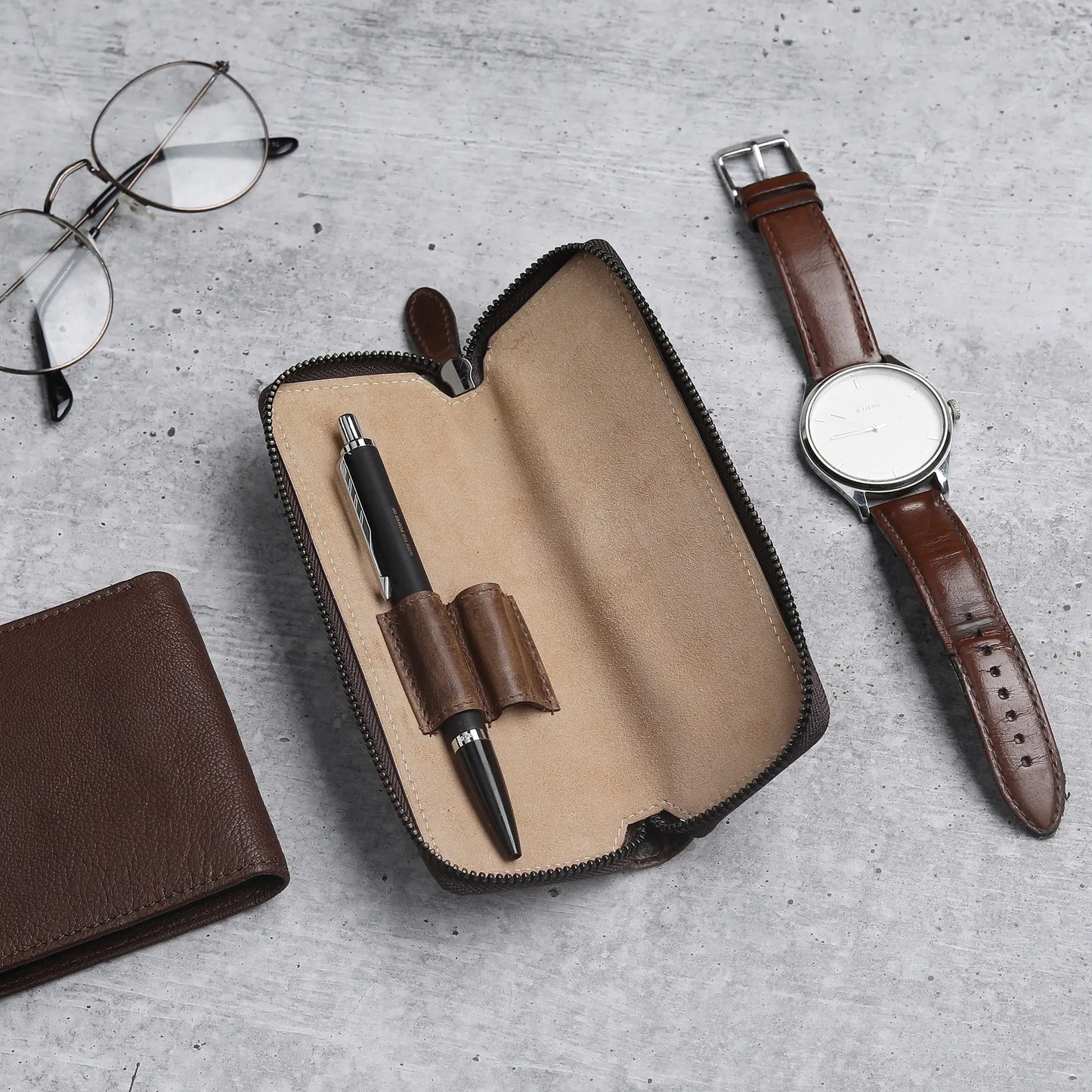 Leather wallet, pen case with pen, glasses, and watch on a gray surface