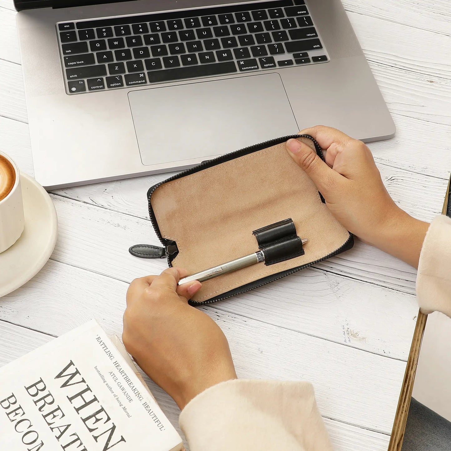 Person holding a brown leather pen case with a pen on a desk next to a laptop and book.
