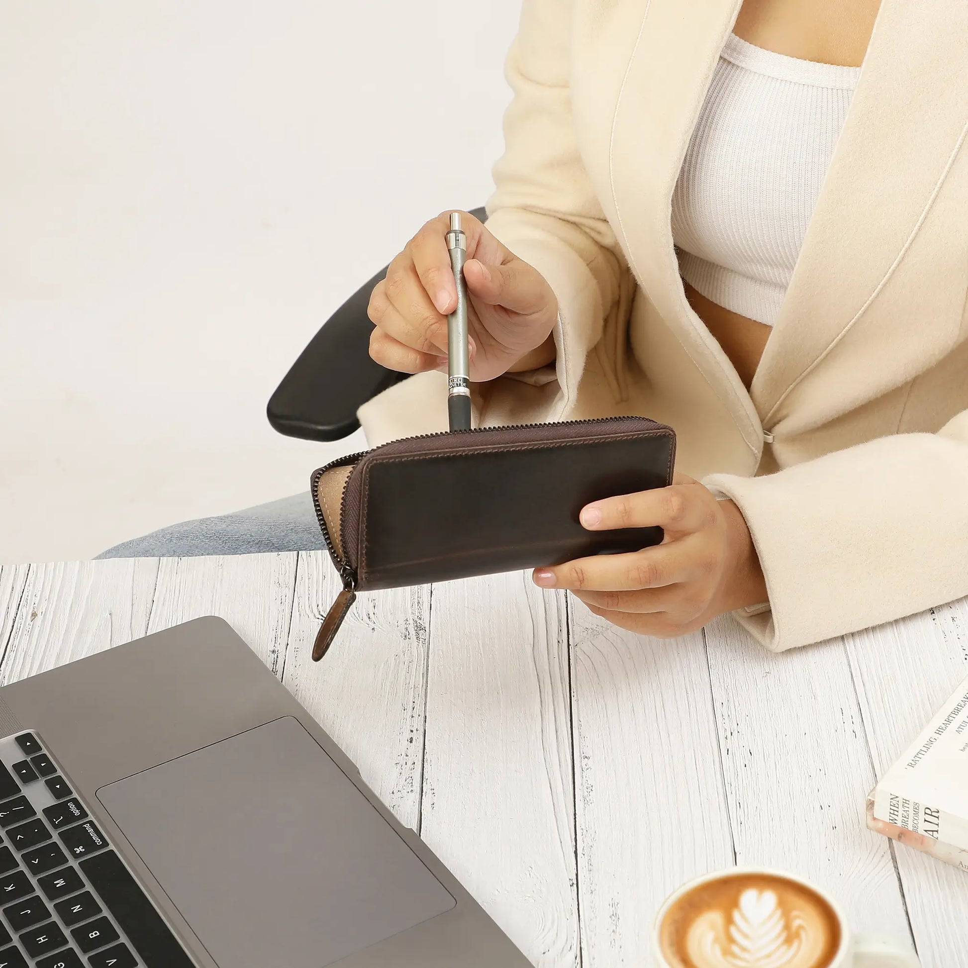 Person holding a pen  and pen case next to a laptop on a wooden surface