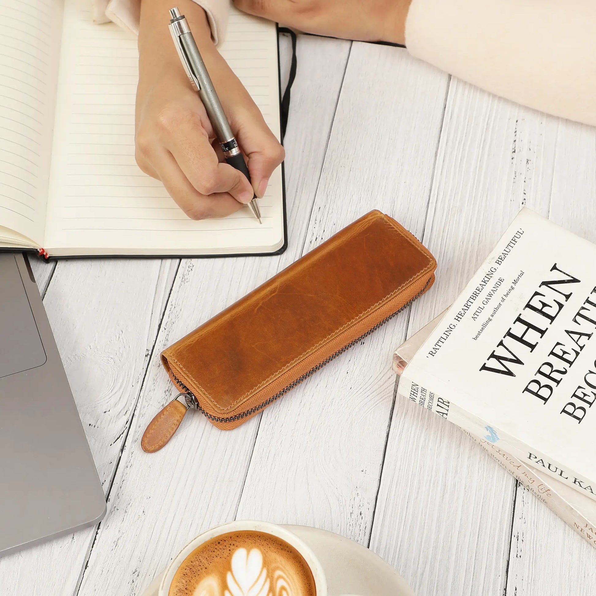 Person writing in a notebook with a tan leather pen case, book, and coffee on a wooden surface.